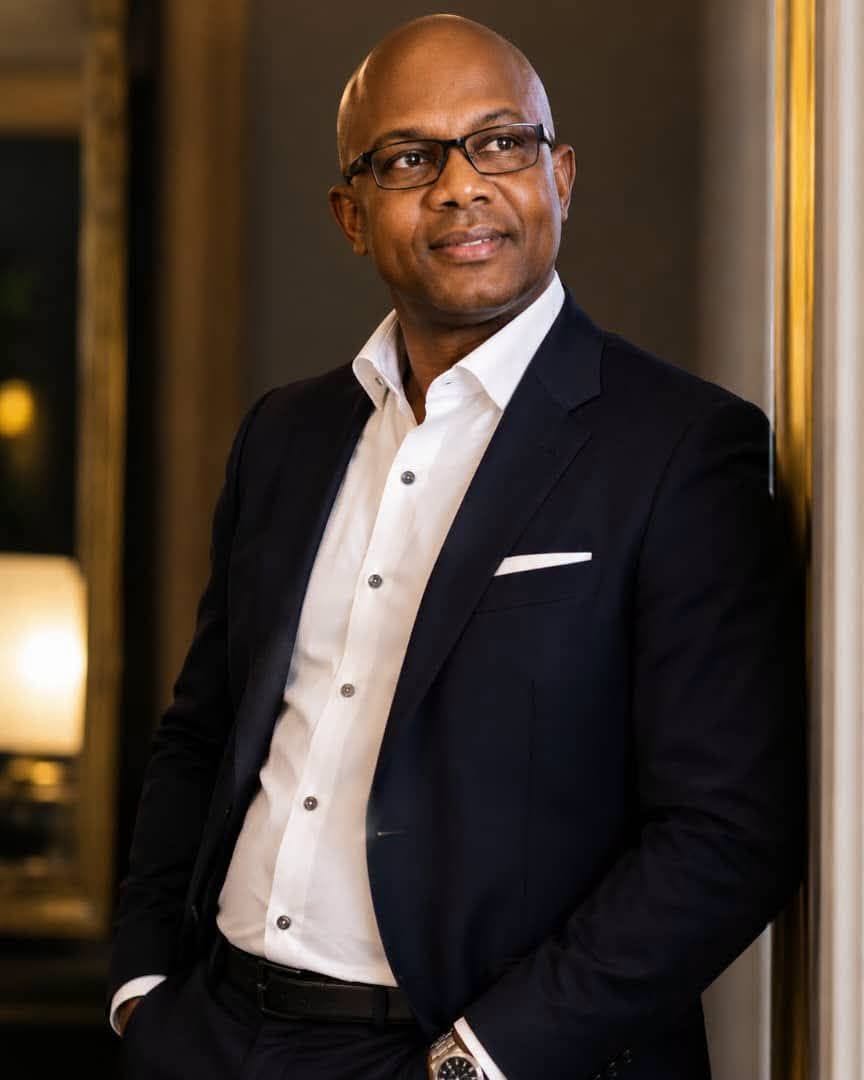 Professional portrait of Tidiane Soumah in a navy blazer, leaning against a pillar in a warmly lit room